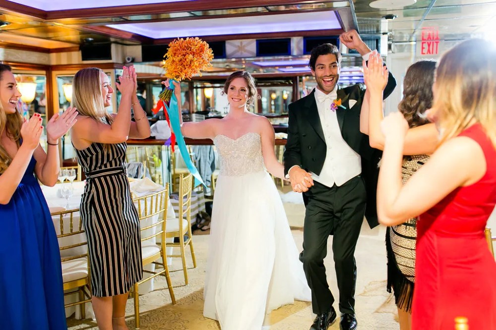 A joyful newlywed couple walking hand in hand as they enter a reception venue on a cruise ship. The bride is in a strapless white gown, and the groom is in a black tuxedo with a bow tie. They are surrounded by cheering guests, clapping and celebrating the couple's entrance. The bride holds a vibrant bouquet of orange flowers with colorful ribbons