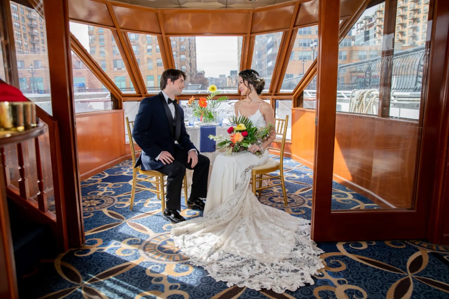 A bride and groom seated together on a yacht, having an intimate moment in a beautifully decorated room with large windows offering views of the city skyline. The bride is wearing an elegant lace wedding dress with a long train, while the groom is in a black tuxedo. They are smiling at each other, with the table in front of them adorned with bright, tropical flowers.