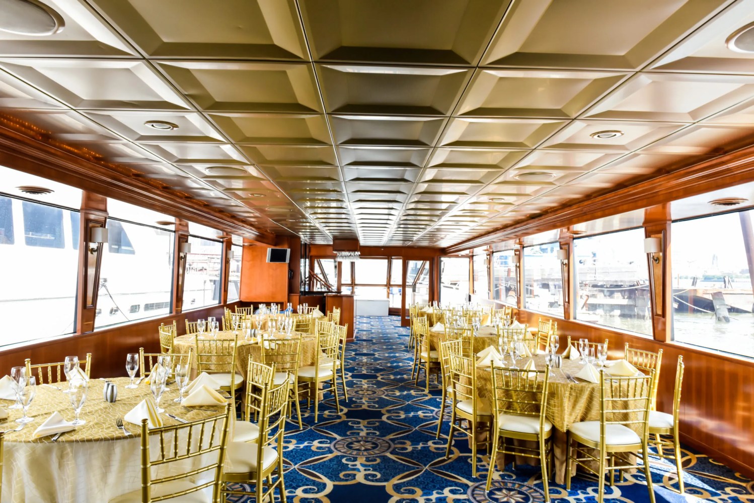 The interior of an elegant dining area on a cruise ship. The room is set up with round tables covered in golden tablecloths, accompanied by gold chairs. Large windows run along both sides of the room, offering views of the outside. The ceiling has a grid-like design with recessed lighting, and the room is decorated with a blue and gold-patterned carpet, creating a luxurious atmosphere.